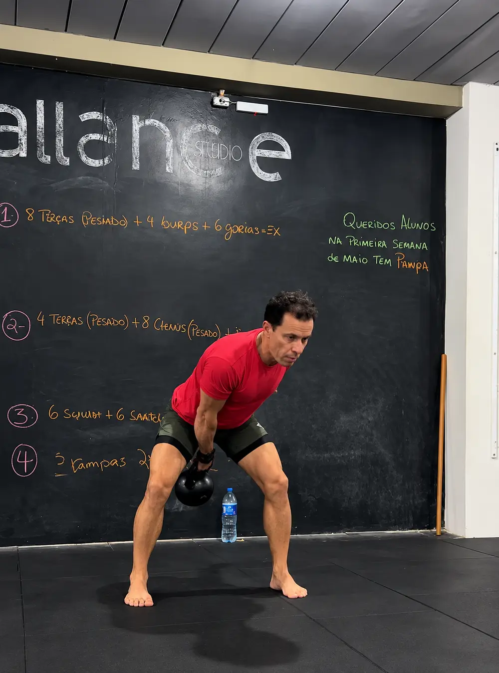Aluno treinando com kettlebell em frente à lousa com o treino do dia